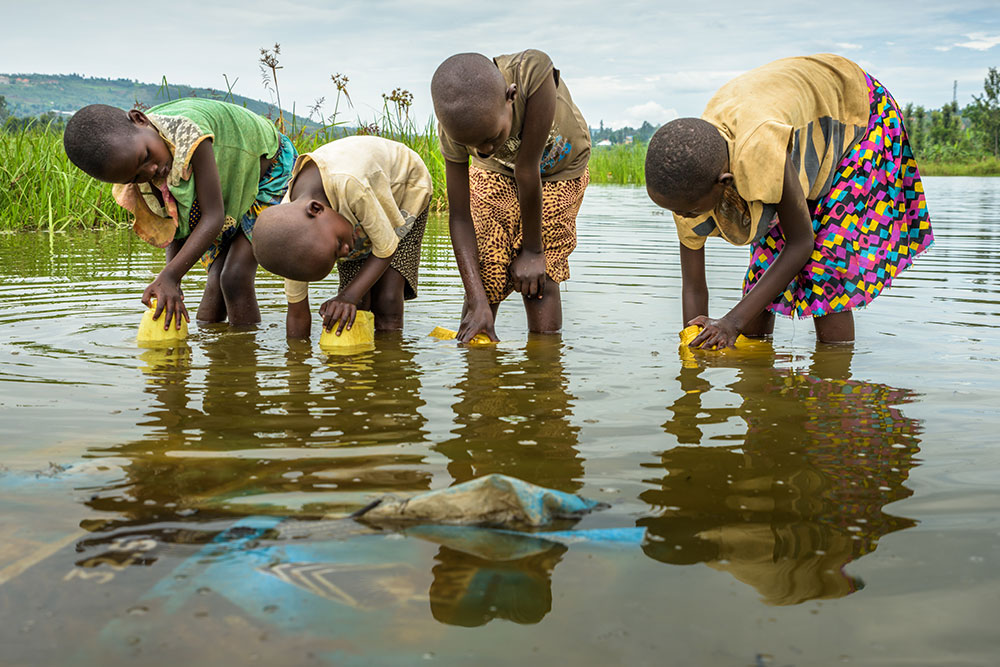 Four young children wade into a lake to fill yellow, plastic bottles with water. Garbage can be seen just under the surface of the lake a short distance from the children.